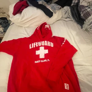 Röd hoodie från Lifeguard - Säljer en cool röd hoodie från Lifeguard med texten 'Lifeguard Marco Island, FL' tryckt på framsidan. Perfekt för en avslappnad stil eller som ett statement-plagg. Den är i bra skick och har en skön passform med en stor huva.