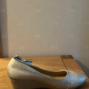 Beige kilklackskor med paljettrosett - Snygga beige kilklackskor i mycket bra skick. De har en glittrig paljettrosett på hälen och en dekorativ paljettkant framtill. Perfekta för festliga tillfällen eller en utekväll. Bekväma och stiliga!