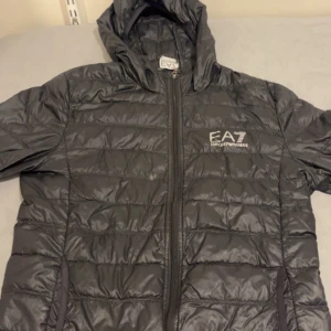 Svart pufferjacka från EA7 Emporio Armani - Säljer en snygg svart pufferjacka från EA7 Emporio Armani. Jackan har en stilren design med dragkedja och huva, perfekt för kyliga dagar. Den är i mycket bra skick och passar både till vardags och lite finare tillfällen. Perfekt för höst och vår!