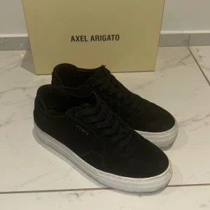 Svarta sneakers från Axel Arigato - Säljer ett par svarta sneakers från Axel Arigato i bra skick. De har en stilren design med svart mocka och vita sulor. Perfekta för både vardag och fest! Skorna har snörning och är bekväma att bära. Kommer med originalkartong.
