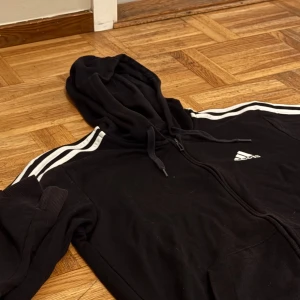 Zip hoodie från Adidas - Säljer en svart hoodie från Adidas i använd skick. Den har de klassiska tre vita ränderna längs ärmarna och en dragkedja framtill. Perfekt för höst och vår, eller när du bara vill ha något bekvämt. Endast 99!