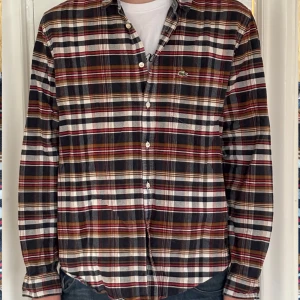 Rutig skjorta från Lacoste - Säljer en snygg rutig skjorta från Lacoste i storlek XXL (men sitter som en M/L). Skjortan har långa ärmar och klassiska knappar framtill. Perfekt för höst och vår med sina jordnära färger i brunt, blått och rött. Den är i mycket bra skick.