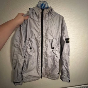 Stone Island windbreaker - Säljer en snygg silvrig jacka från Stone Island. Den har en praktisk dragkedja och två stora fickor med knappar framtill. Perfekt för höst och vår, och den har långa ärmar. Jackan är i bra skick och passar perfekt för en utåtstickande outfit! Jackan tål vind och vatten och är väldigt praktisk samt otroligt snygg. Den är i fint skick och fler bilder kan skickas om det önskas!