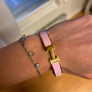 Guld och rosa armband - Säljer ett stilrent armband i guld med rosa detaljer. Perfekt för att ge en färgklick till din outfit. Armbandet har en elegant design med ett H-format spänne. Passar både till vardags och fest!