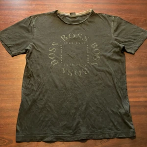 Svart t-shirt från Hugo Boss {S} - Säljer en svart t-shirt från Hugo Boss i storlek S. Den har ett stilrent tryck med 'BOSS' i en cirkel på framsidan. T-shirten är i bra skick och har inga hål eller fläckar. 