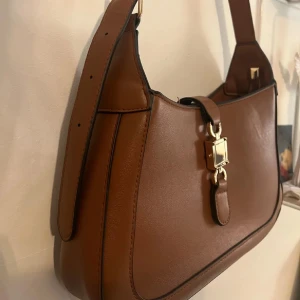 Brun axelväska med gulddetaljer - Snygg brun axelväska i skinn med eleganta gulddetaljer. Perfekt för både vardag och fest. Väskan har en justerbar axelrem och en stilren design som passar till alla outfits. Köpt här på plick, den är från glitter och är i mycket bra skick 💕