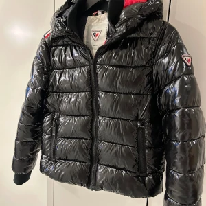 Svart glansig dunjacka från Rossignol - Dunjacka junior strl 10  från Rossignol. Insidan är fodrad med rött, vitt och svart tyg. Blank svart utsida. Perfekt för både kille och tjej! Knappt använd. 