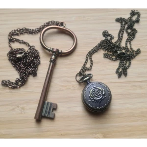 Klocka, nyckel halsband brons/kopparfärgade - Okända material. Osäker på om det går att få klockan att fungera, vet inte hur man öppnar den för att se. Klocka: kedja: 80, vidd: 2,7, höjd: 4. Nyckel: vkedja: 77, vidd: 2,6, höjd: 9,5.