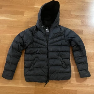 Svart dunjacka från Nike - Säljer en svart puffer jacka från Nike i bra skick. Jackan har en skön huva och dragkedja framtill. Perfekt för kalla dagar med sin pufferdesign som håller värmen. Den har praktiska fickor med knappar. Passar både till vardags och sportiga aktiviteter. 13-15 år