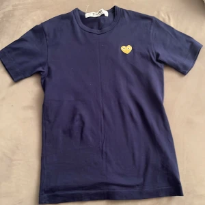 Mörkblå t-shirt från Comme des Garçons - Säljer en snygg mörkblå t-shirt från Comme des Garçons med det ikoniska hjärtemärket på bröstet. T-shirten är i bomull och har en normal passform med korta ärmar. Perfekt för en stilren och avslappnad look!