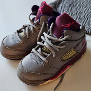 Baby Air Jordan ,strlk EU 22 - Säljer ett par grå och vinröda Nike Air Jordans i barnstorlek. Skorna har snörning och en cool design med Jumpman-loggan i rosa. Perfekta för små sneakerheads som vill ha stil! Använda men i bra skick.