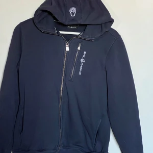 Marinblå huvtröja från Sail Racing - Säljer en marinblå huvtröja från Sail Racing i mycket bra skick. Den har en dragkedja framtill och en praktisk bröstficka med dragkedja. Perfekt för höst och vår, med en stilren design och bekväm passform. Den är storlek 170 så skulle säga runt small/medium