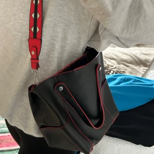 Svart axelväska med röda detaljer - Snygg svart axelväska med röda sömmar och detaljer. Väskan har en axelrem med ett mönster i rött, svart och vitt. Den har en magnetknapp för enkel öppning och stängning.