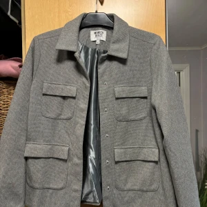 Grå kavaj från Monica Geuze x NA-KD - Säljer en stilren grå kavaj från Monica Geuze x NA-KD. Den har en klassisk design med fyra fickor framtill och en diskret rutigt mönster. Perfekt för både vardag och fest! 🖤