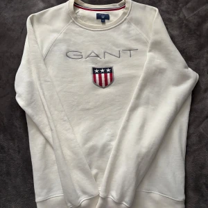 S Vit sweatshirt från GANT - Säljer en snygg vit sweatshirt från GANT med broderad logga och sköld på bröstet. Tröjan har långa ärmar och är perfekt för en avslappnad stil. Passar både till vardags och lite finare tillfällen. Superbekväm och stilren!
