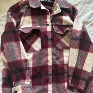 Rutig kappa från Zara - Säljer en superfin rutig Kappa från Zara i storlek S. Den har en mysig och varm känsla med långa ärmar och knappar framtill. Perfekt för höst och vår! Färgerna är en mix av vinröd, vit och grå. Passar både till vardags och lite mer uppklädda tillfällen.