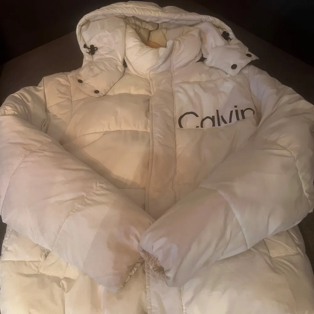 Säljer en snygg vit pufferjacka från Calvin Klein. Jackan är perfekt för kalla dagar med sin tjocka vaddering och har en stor huva för extra värme. Den har långa ärmar och en dragkedja framtill. Stort Calvin Klein-tryck på både fram- och baksidan ger en cool look. Perfekt för vintern!. Jackor.