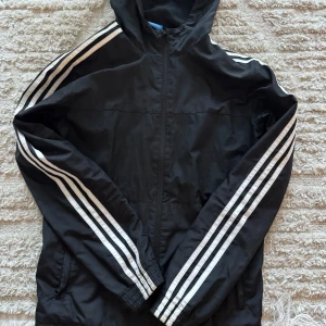 Svart vindjacka från Adidas - Säljer en svart vindjacka från Adidas med de klassiska tre vita ränderna längs ärmarna. Jackan har en dragkedja framtill och är perfekt för sportiga aktiviteter eller som en lätt jacka till vardags. 