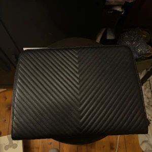 Svart quiltad datorväska - Säljer en stilren svart datorväska med quiltat mönster. Väskan har en dragkedja upptill och är perfekt för att skydda din laptop. Den är i syntetmaterial och har en enkel men elegant design. Perfekt för både vardag och arbete!