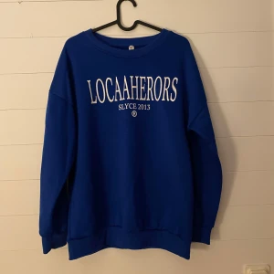 Blå sweatshirt med tryck - Säljer en snygg blå sweatshirt med texten 'LOCAAHERORS SLYCE 2013' tryckt på framsidan. Tröjan har en avslappnad passform och är perfekt för både vardag och chill. Den är långärmad och gjord i ett mjukt material som känns skönt mot huden. Perfekt för höst och vinter!
