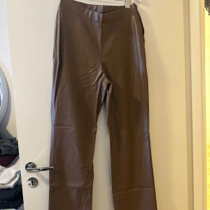 Bruna skinnbyxor från Only - Säljer ett par snygga bruna skinnbyxor från NLY Trend. De är högmidjade och har en stilren design som passar perfekt för både vardag och fest. Byxorna är i syntetmaterial och har en glansig finish.