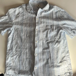 Ljusblå och vit randig skjorta från H&M - Säljer en snygg ljusblå och vit randig skjorta från H&M i storlek M. Skjortan är kortärmad och har en bröstficka. Den är tillverkad av 52% linne och 48% bomull, vilket gör den perfekt för varma sommardagar eller fester. Endast använd 1 gång.