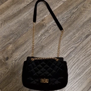 Svart quiltad axelväska med guldkedja - Snygg svart axelväska i quiltad design med guldkedja och vridlås. En mobil får lätt plats tillsammans med flera saker. På sista bilden kan ni jämföra storleken med en hand