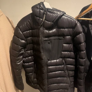 Svart dunjacka från C.P. Company - Säljer en svart dunjacka från C.P. Company med huva och inbyggda glasögon. Jackan har en quiltad design och dragkedja framtill. Perfekt för kalla dagar. Säljes då den blivit för stor för mig. Nypris runt 6000 lite osäker då jag har haft den länge och det är en äldre modell. Jackan är i fint skick då jag inte använt den mer än drygt en vinter👍🏻|
