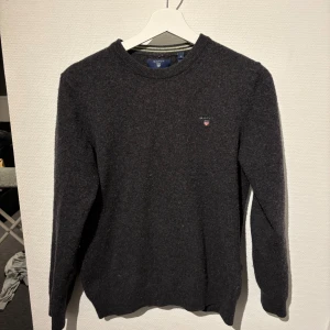 Mörkgrå ulltröja GANT - Säljer en stilren mörkgrå tröja från GANT i superfin lammull. Tröjan har en klassisk rund halsringning och ett diskret broderat märke på bröstet. Perfekt för kyliga dagar! Liten i storlek pga tvättad, passar S och XS