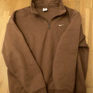 Brun tröja med dragkedja från Nike - Säljer en brun tröja från Nike med halv dragkedja och det klassiska Nike-logotypen på bröstet. Perfekt för en avslappnad stil. Obs!!! Tröjan är oversized!