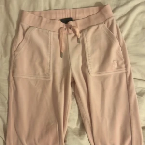 Rosa mjukisbyxor från Juicy Couture - Säljer ett par ljusrosa mjukisbyxor från Juicy Couture. Byxorna har en bekväm resårmidja med snörning och praktiska fickor både fram och bak. Detaljerad med Juicy Coutures logga broderad på framsidan. Perfekta för en avslappnad stil.