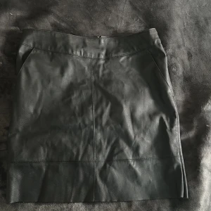 Svart skinnkjol med dragkedja - Snygg svart skinnkjol med en stilren design. Kjolen har en dragkedja baktill och en kort längd som ger en modern look. Perfekt för att addera lite edge till din outfit.