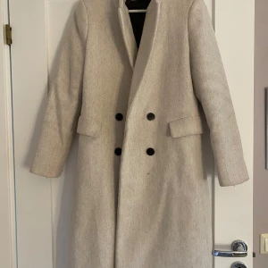 Beige kappa från MNG - Säljer en elegant beige kappa från MNG i storlek S. Den har en klassisk dubbelknäppt design med svarta knappar och långa ärmar. Perfekt för höst och vår, och ger en stilren look till vilken outfit som helst. Kappan har ett hål i innetyget men som lätt går att sy ihop om man skulle vilja det, och är lite smått nopprig, annars bra skick!💓💓