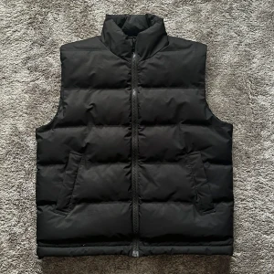 Svart dunväst från Weekday - Säljer en stilren svart dunväst från Weekday i storlek M. Västen har en dragkedja framtill och två praktiska fickor. Perfekt för höst och vinter när du vill hålla värmen utan att kompromissa med stilen. Passar både till vardags och mer avslappnade tillfällen. 