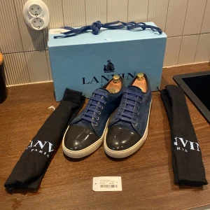 Blå sneakers från Lanvin - Säljer ett par snygga blå sneakers från Lanvin i mycket bra skick. Skorna har en cool design med blå mocka och svart glans på tån. De kommer med originalförpackning och två svarta skopåsar. Perfekta för både vardag och fest! Det tillkommer samt ett litet ” kvitto” om man vill säga så. Allt OG tillkommer samt