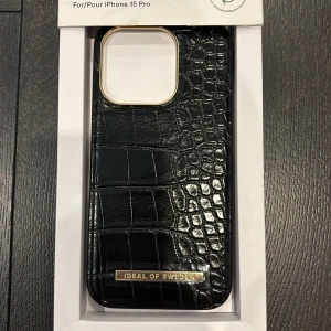 Svart mobilskal från Ideal of Sweden - Snyggt svart mobilskal för iPhone 15 Pro från Ideal of Sweden. Skalet har en elegant krokodilstruktur och en stilren design med gulddetaljer runt kameran och logotypen. Perfekt för att ge din telefon ett lyxigt utseende samtidigt som den skyddas. Den är helt ny aldrig andvänd. Original pris 499kr 