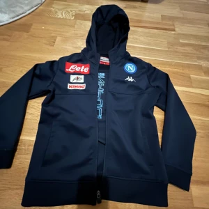 Marinblå set, Napoli - Säljer en mörkblå huvtröja från Kappa med dragkedja och flera logotyper, inklusive Napoli och Lete. Perfekt för unga sportfantaster. Tröjan har långa ärmar och en praktisk huva.
