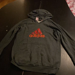 Mörkgrå hoodie från Adidas - Säljer en mörkgrå hoodie från Adidas med röd logga på bröstet. Tröjan har en klassisk känguruficka och huva. Perfekt för en avslappnad stil.