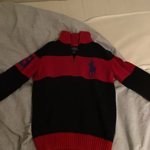Svart och röd tröja från Ralph Lauren - Snygg svart och röd tröja från Ralph Lauren med en stor logga på bröstet och siffran 3 på ärmen. Tröjan har en dragkedja vid halsen och är stickad för extra komfort. Perfekt för kyligare dagar!