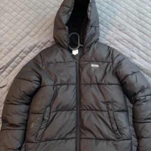 Svart pufferjacka från Boss - Snygg svart pufferjacka från Boss med dragkedja och huva. Perfekt för kyligare dagar med sin quiltade design som håller värmen. Jackan har en stilren look med diskret logga på bröstet. Två små knappt märkbara hål. Storlek passar XS. Pris kan diskuteras 