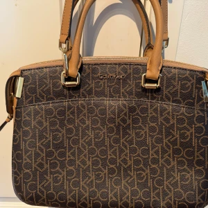 Brun axelväska från Calvin Klein - Snygg brun axelväska från Calvin Klein med monogrammönster. Väskan har justerbara axelremmar och handtag i beige. Perfekt för att bära dina dagliga nödvändigheter med stil.