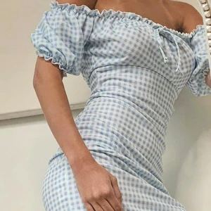 Blåvit rutig klänning - Charmig blåvit rutig klänning med offshoulder-design och korta puffärmar. Klänningen har en smickrande passform med volangdetaljer vid halsen och ärmarna. Perfekt för somriga dagar. St 36/38