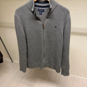 Grå stickad tröja från Polo Ralph Lauren - Säljer en stilren grå stickad tröja från Polo Ralph Lauren med dragkedja och broderad logotyp på bröstet. Tröjan har en hög krage och ribbade muddar för extra komfort. Perfekt för kyliga dagar! Storlek XL i barn så passar S i vuxen