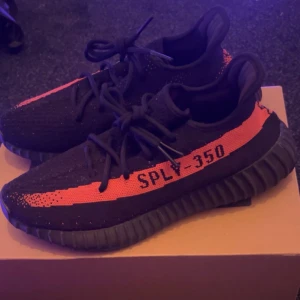 Adidas Yeezy Boost 350 V2 - Snygga Adidas Yeezy Boost 350 V2 sneakers använda 1 gång i svart med en iögonfallande röd rand och texten 'SPLY-350'. Skorna har en stickad ovandel och en bekväm, ribbad sula. Perfekta för dig som vill ha en stilren och modern look.