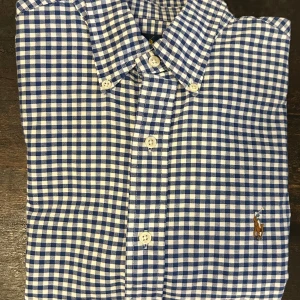 Blå och vit rutig skjorta från Ralph Lauren stl S - Säljer en snygg blå och vit rutig skjorta från Ralph Lauren i storlek S. Skjortan är slim fit och har långa ärmar med knappar framtill. Den är broderad med det klassiska Ralph Lauren-logotypen på bröstet. Perfekt för både vardag och fest! 100% bomull