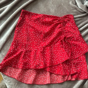 Röd prickig kjol med volang - Superfin röd kjol med vita prickar och volangdetaljer. Den har en snygg omlott-look och en knytning framtill som ger en extra touch. Perfekt för sommarens fester eller en dag på stan. Kjolen är kort och har en härlig flowy känsla. Passar perfekt med en enkel topp och sneakers eller klackar för en mer uppklädd stil.