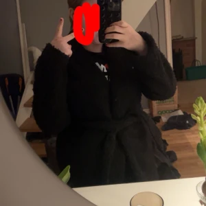 Svart teddyjacka med knyt - Mysig svart jacka med knyt i midjan. Jackan har långa ärmar och en mjuk textur. På insidan är det silkes materixal och utsidan lurvigare. Jackan är ganska använd. Den är mellan-lång och går ner 10cm under rumpan