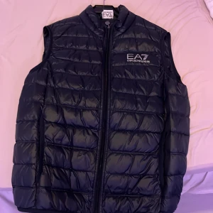 Svart dunväst från Emporio Armani - Snygg svart dunväst från Emporio Armani med dragkedja framtill. Västen har en quiltad design och logotyp på bröstet. Perfekt för kyligare dagar.