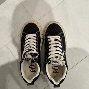 Svarta sneakers från Eytys - Snygga svarta sneakers från Eytys med vit sula och snörning. Skorna har en unik, grov sula och lila dragflik baktill. Perfekta för en stilren look. ÄLSKAR DE men tyvärr är de för stora:(