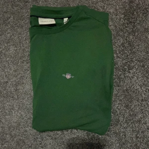 Grön t-shirt från GANT - Säljer en snygg grön t-shirt från GANT i storlek S. Den har en liten broderad logga på bröstet som ger en stilren look. Perfekt för vardagsbruk eller en avslappnad dag ute. Passar bra till jeans eller shorts! (Accepterar endast Swish pga lurendrejerier) (Checka gärna in kläder på min profil och köp tillsammans till ett rabatterat pris)
